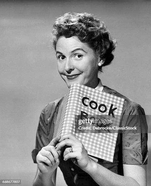 1950s SMILING WOMAN LOOKING AT CAMERA OVER TOP OF A COOK BOOK