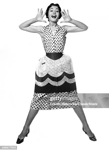 1950s WOMAN IN DRESS & APRON JUMPING IN AIR LEGS APART & PALMS SPREAD TO EITHER SIDE OF FACE WITH MOUTH OPEN LOOKING AT CAMERA