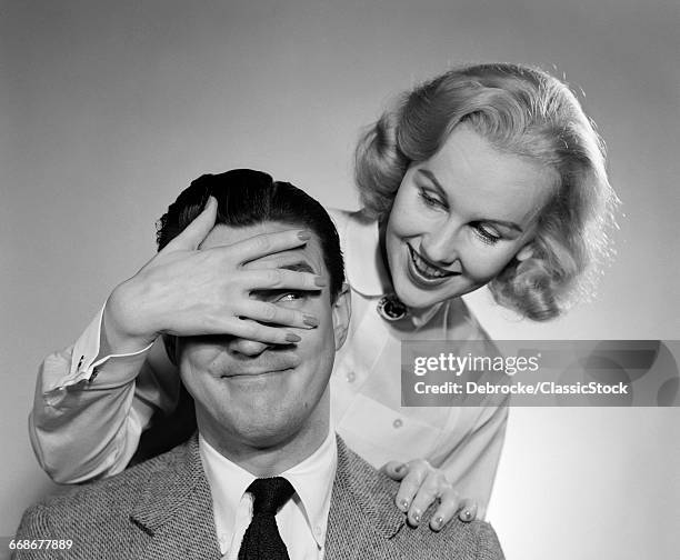 1950s WOMAN STANDING BEHIND MAN WITH HAND OVER HIS EYES