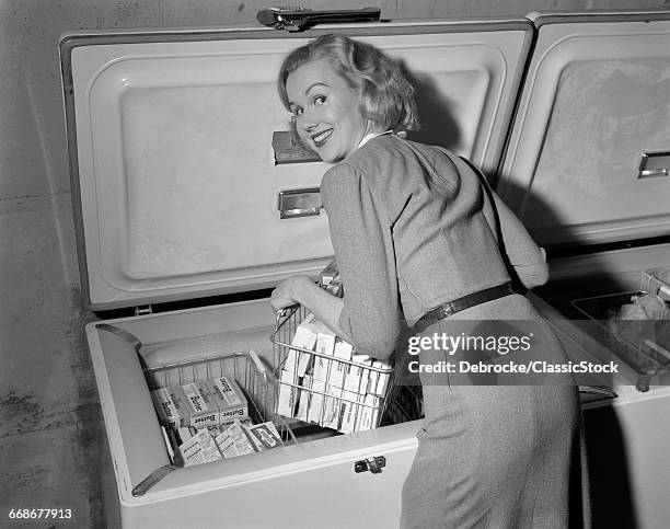 1950s BLOND WOMAN LIFTING WIRE BASKET OF FOOD ITEMS FROM A DEEP FREEZER LOOKING AT CAMERA OVER HER SHOULDER