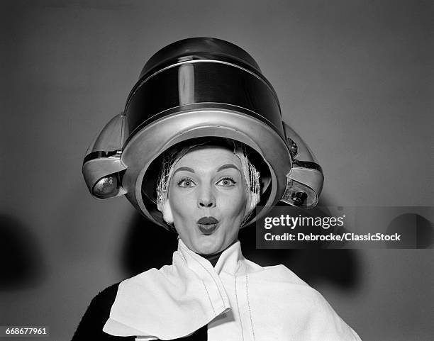 1950s WOMAN UNDER HAIR DRYER WITH TOWEL ON SHOULDERS AND HAIR NET LOOKING AT CAMERA