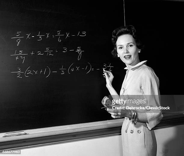 1950s MATH TEACHER STANDING IN FRONT OF BOARD HOLDING CHALK AND ERASER LOOKING AT CAMERA