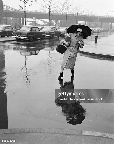 1950s WOMAN IN RAIN COAT HAT BOOTS HOLDING UMBRELLA HANDBAG STEPPING TO AVOID PUDDLES WHILE CROSSING CITY STREET IN FOUL WEATHER