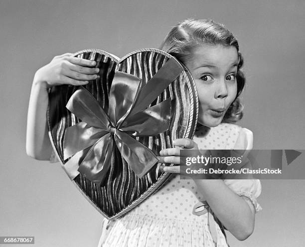 1950s SURPRISED LITTLE GIRL LOOKING AT CAMERA FROM BEHIND BIG HEART SHAPED BOX OF VALENTINE CANDY
