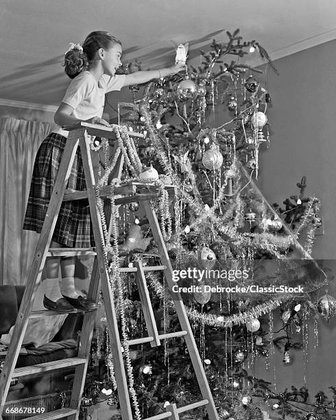 1950s JUVENILE GIRL ON LADDER TRYING TO PUT ANGEL ON TOP OF CHRISTMAS TREE