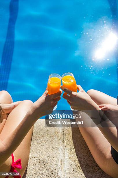 two women sitting on the edge of a pool toasting with glasses of orange juice, partial view - friends poolside stock pictures, royalty-free photos & images