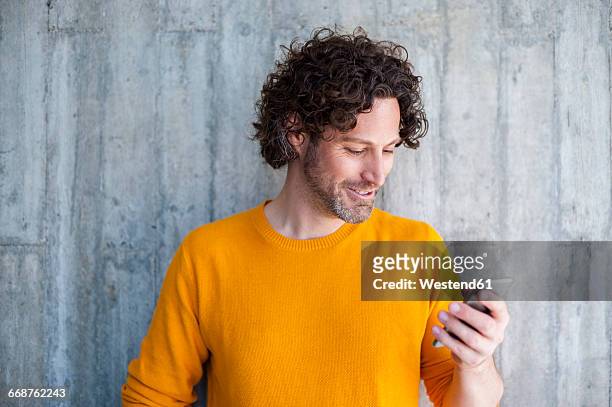 smiling man with curly brown hair looking on his smartphone - portrait mauer stock-fotos und bilder
