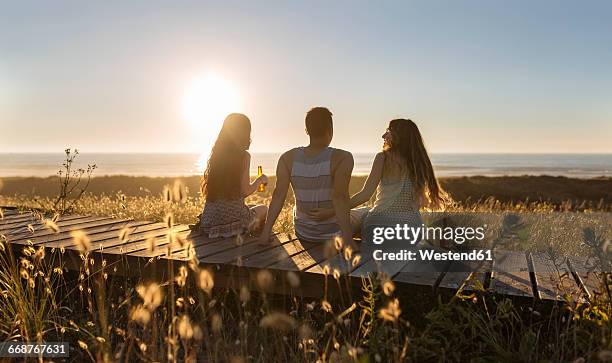 friends sitting on boardwalk at the beach, drinking beer at sunset - men friends beer outside stock-fotos und bilder