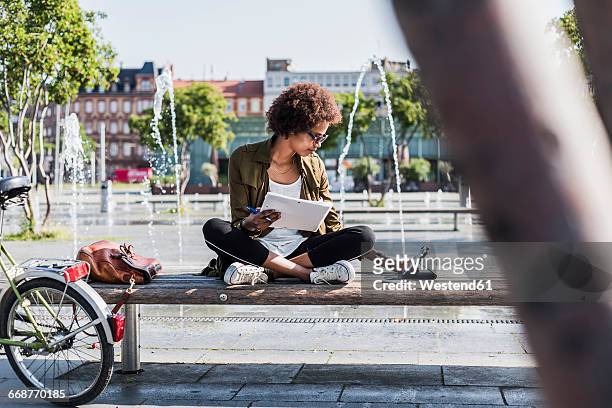 young woman sitting on a bench with notebbok looking at digital tablet - werk laptop buiten stockfoto's en -beelden