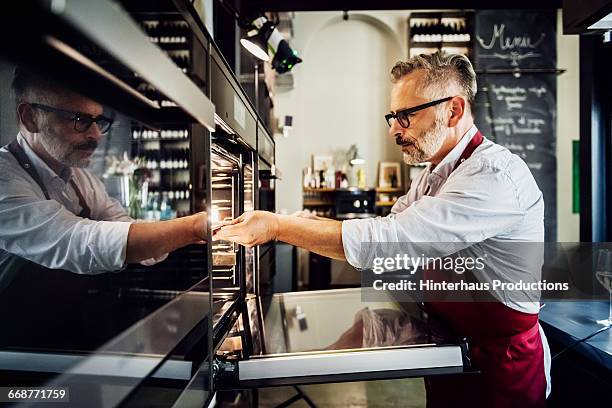 mature man putting food inside an oven - kochschürze stock-fotos und bilder