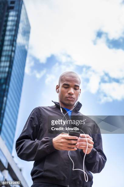 hombre de raza mixta en ropa deportiva ajusta mp3 reproductor de música al aire libre entre los rascacielos de oficinas soleadas - black pants fotografías e imágenes de stock