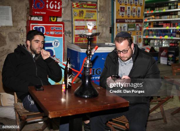 At a pub in the Nahalat Shiv'a neighborhood, a pair of young men, both with yarmulkes on their heads, sit at a table with bottles of beer before them...