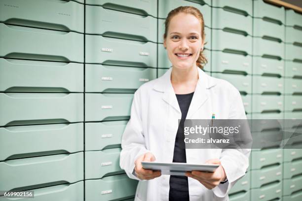 pharmacist holding digital tablet in storage room - female pharmacist with a digital tablet imagens e fotografias de stock