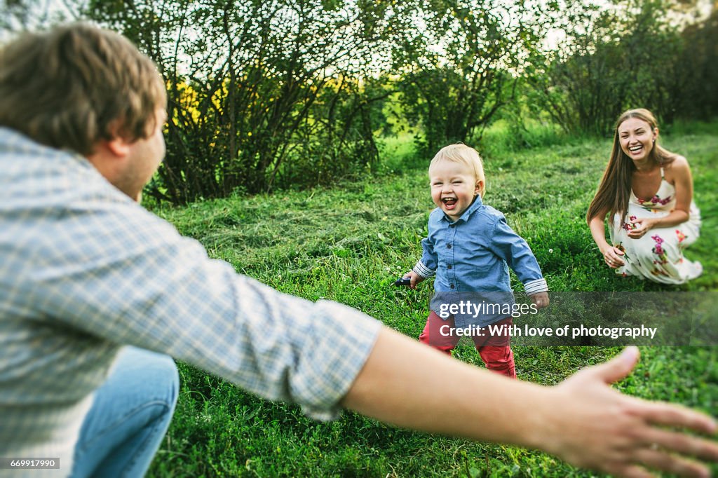 Two parents with little toddler son 1 year old playing outdoors on a sunny day in countryside on green grass