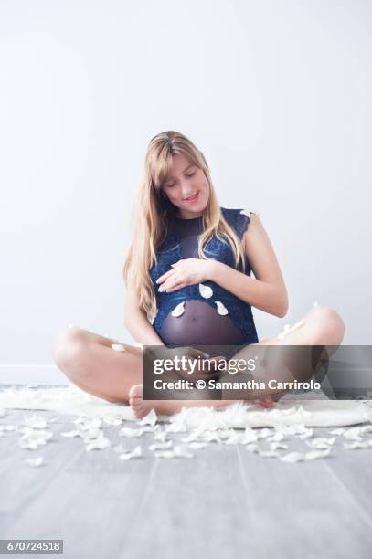 donna incinta seduta su un tappeto bianco. mano sul pancione. intimo / body blu. petali bianchi per terra e in aria. - femminilità stock-fotos und bilder