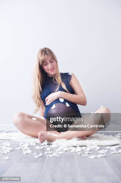 donna incinta seduta su un tappeto bianco. mano sul pancione. intimo / body blu. petali bianchi per terra e in aria. - caucasico stock-fotos und bilder