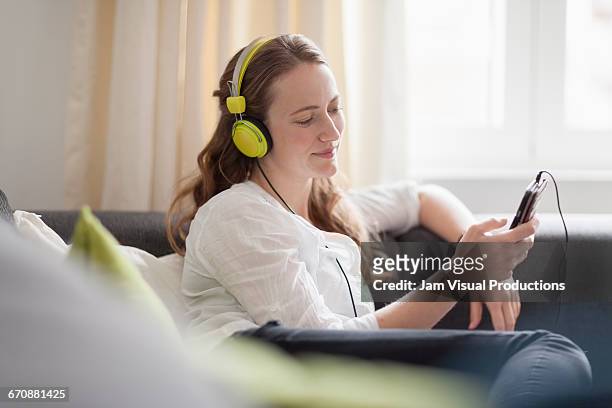 mid-adult woman lying on sofa and listening to music from smart phone - escapismo imagens e fotografias de stock