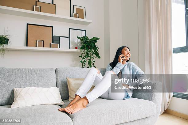 laughing young woman sitting on couch at home telephoning with cell phone - beautiful asian legs - fotografias e filmes do acervo