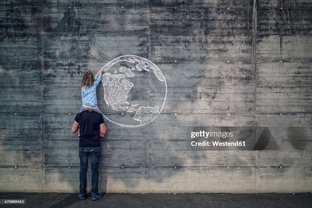 Austria, Salzburg, Father with daughter on his shoulders, the daughter draws with chalk the earth on a concrete wall