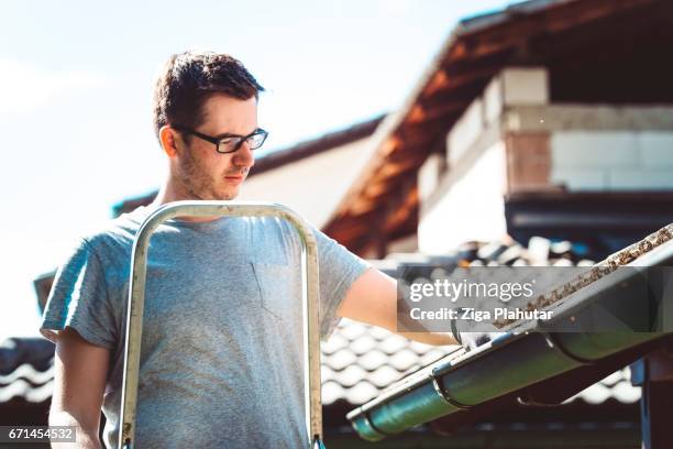 man clearing leaves from gutter up high on ladder - roof gutter stock pictures, royalty-free photos & images