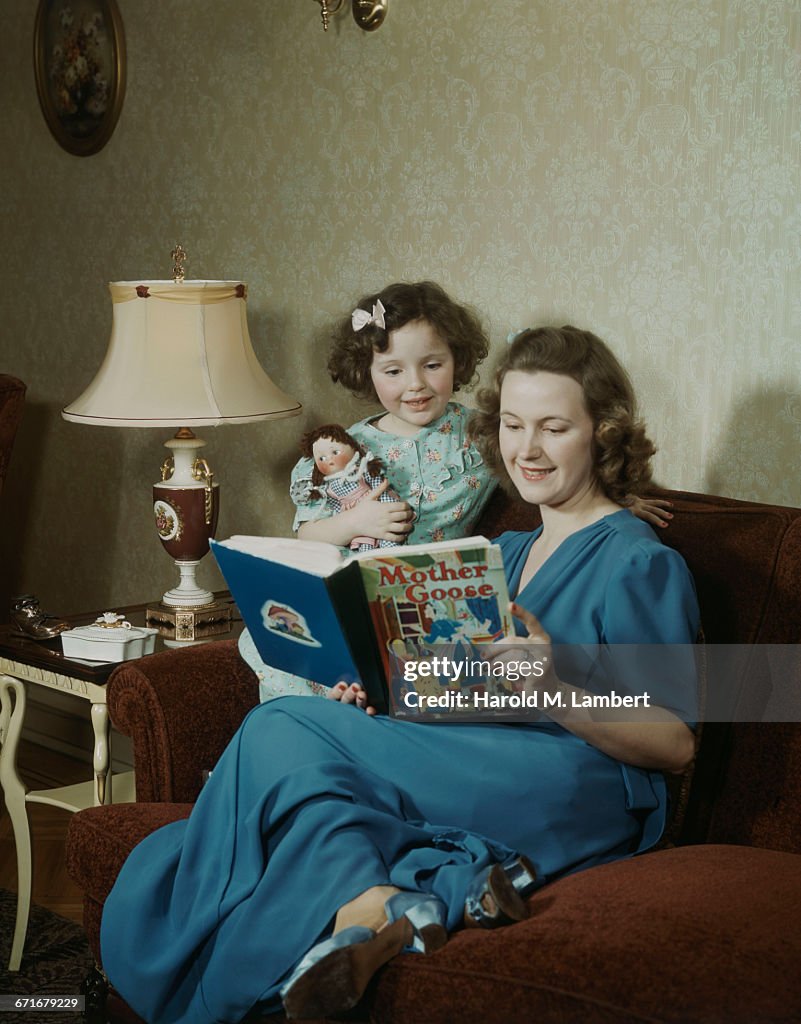 Mother With Daughter Reading Book And Sitting In Sofa 