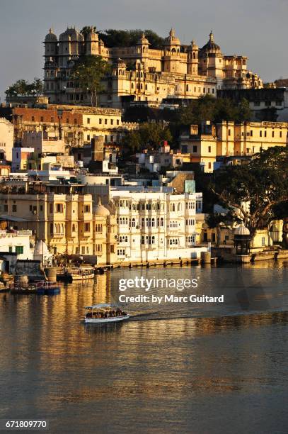 a tour boat drifts beneath the imposing city palace of udaipur at sunset, rajasthan, india - udaipur palace stock-fotos und bilder