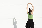pretty girl standing with her back turned and hands up, doing exercises, stretches and relaxes in front of a white wall with space for text