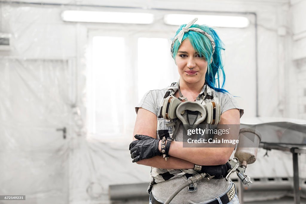 Portrait confident young woman with blue hair with paint gun in auto body shop