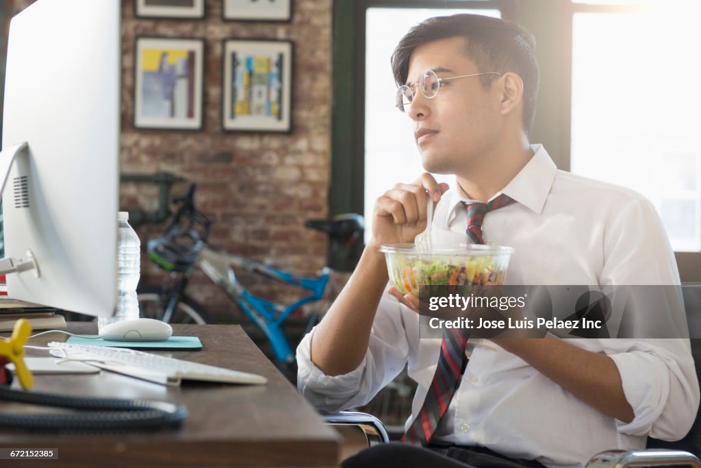 Mixed Race businessman using computer and eating salad at desk