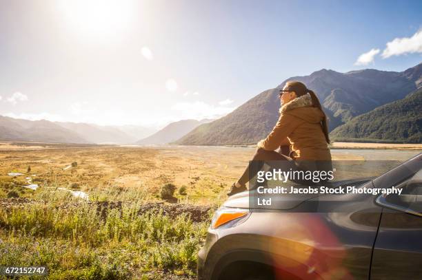 hispanic woman sitting on hood of car admiring scenic view - new zealand holiday stock pictures, royalty-free photos & images