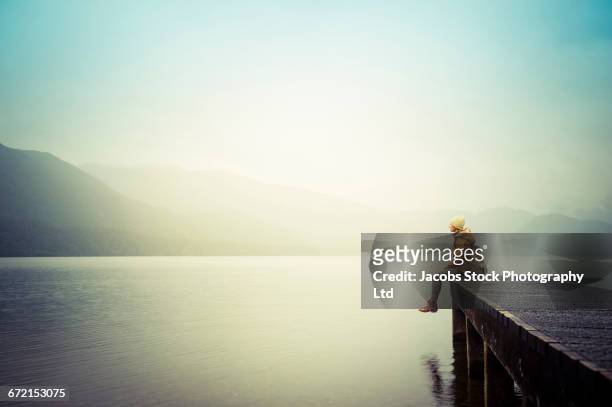 distant hispanic woman wearing coat sitting on dock at mountain lake - fern stock-fotos und bilder
