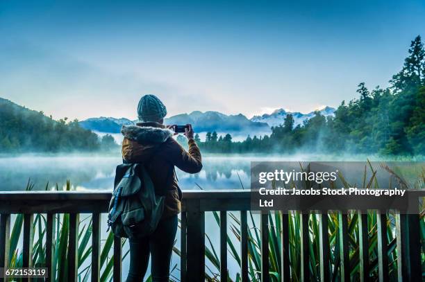 hispanic woman wearing coat photographing foggy mountain lake - eisstockschießen stock-fotos und bilder