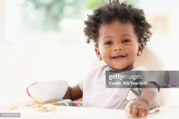 smiling black baby girl eating cereal from bowl in high chair - よだれ掛け ストックフォトと画像