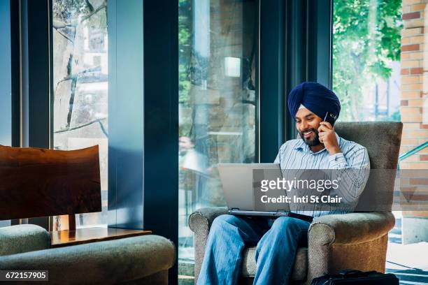 man wearing turban using laptop and cell phone in armchair - sikh foto e immagini stock