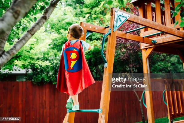 caucasian boy wearing superhero costume climbing on backyard playground - p stock-fotos und bilder