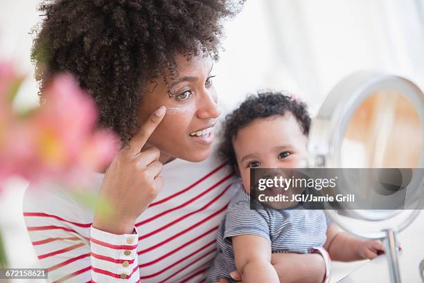 mother holding baby son applying face cream in mirror - baby suncream stockfoto's en -beelden