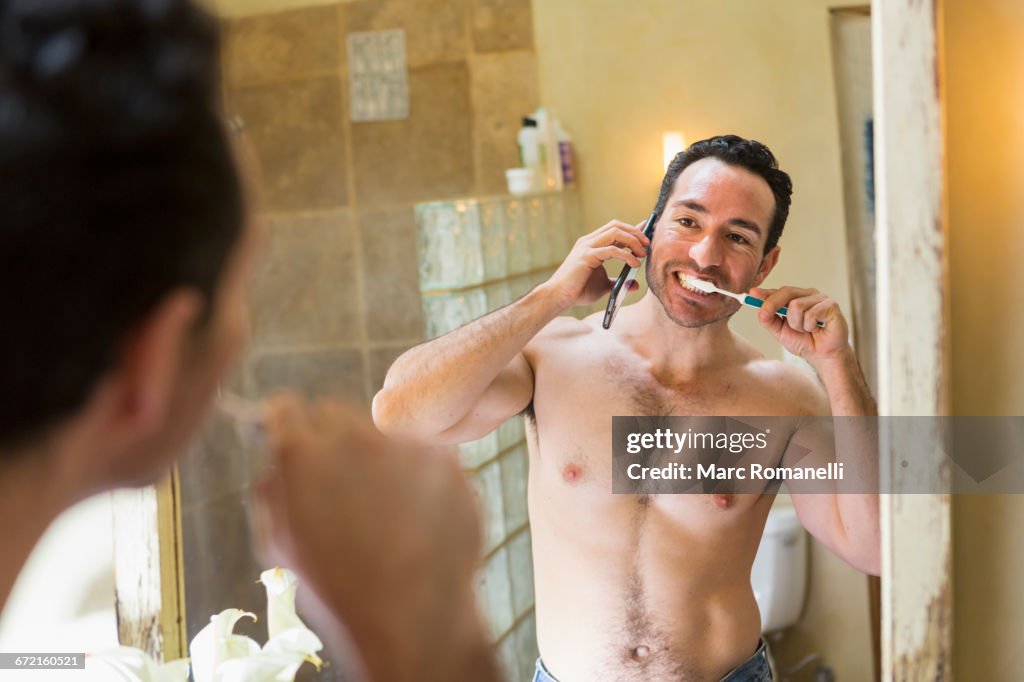 Hispanic man using cell phone and brushing teeth in mirror