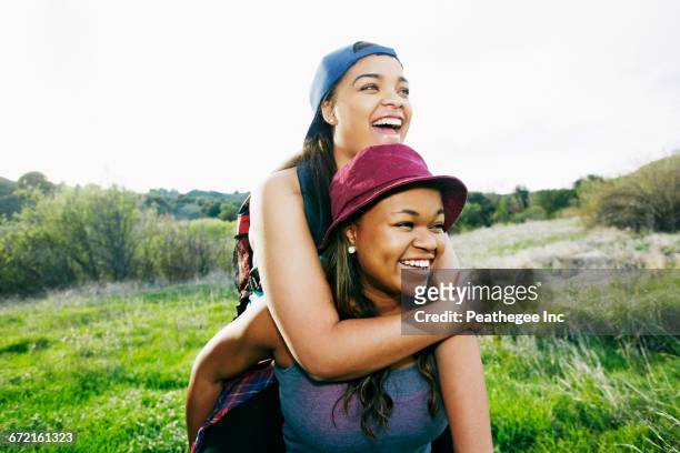 mixed race teenager giving sister piggyback ride in field - african american hiking stock-fotos und bilder