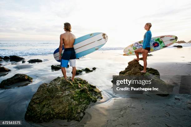 caucasian couple standing on rocks carrying surfboards at beach - malibu stock pictures, royalty-free photos & images