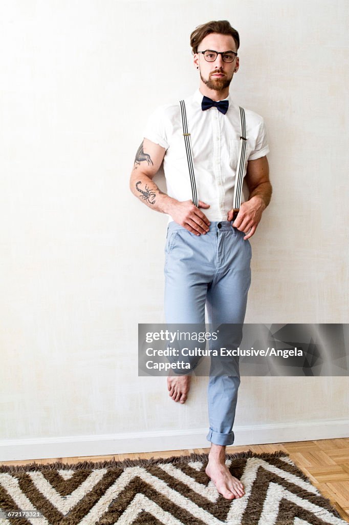 Portrait of cool young man wearing trouser braces and bow tie leaning against wall