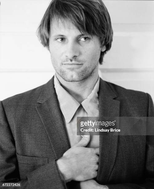 View of Viggo Mortensen standing wearing a button-down shirt and sport coat, he tucks his hands inside his coat lapels, his right hand above his...