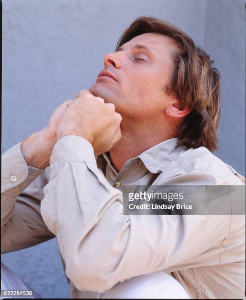Viggo Mortensen in profile, dressed in a tan button-down shirt as he sits on a porch before a blue wall and tilts his head backward, he supports his...
