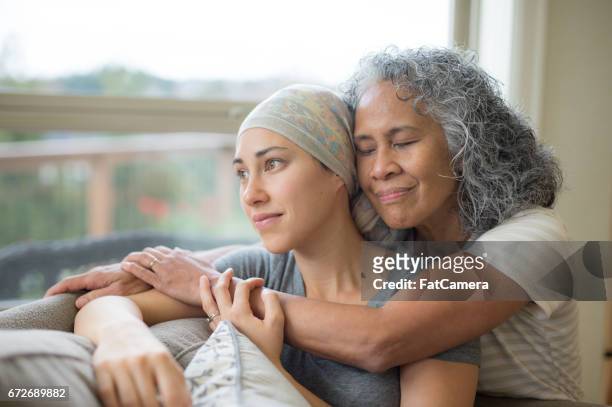 hawaiian woman in 50s embracing her mid-20s daughter on couch who is fighting cancer - cancer illness stock pictures, royalty-free photos & images