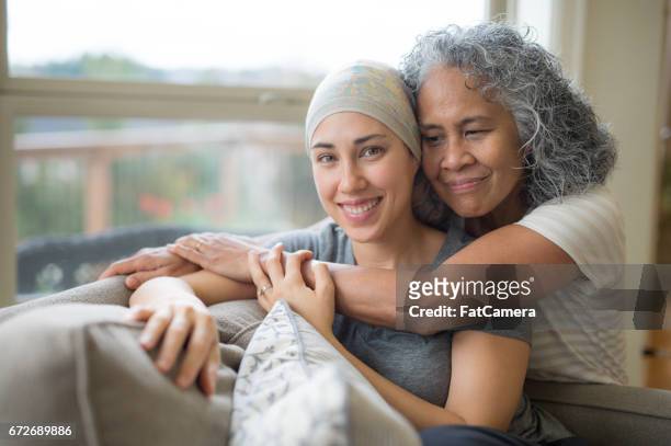 hawaiian woman in 50s embracing her mid-20s daughter on couch who is fighting cancer - cancer 2016 stock pictures, royalty-free photos & images