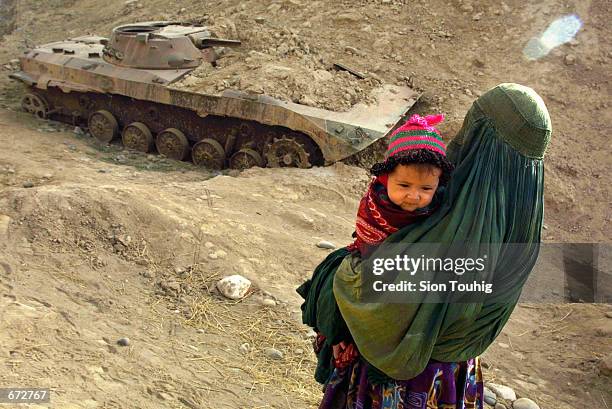 An Afghan refugee woman, clad in a traditional burqa, holds her child and passes a destroyed and abandoned Soviet tank November 21, 2001 as she and...