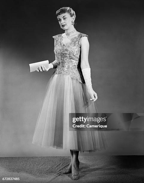 1950s WOMAN STANDING INSIDE WEARING FORMAL EVENING DRESS HOLDING POCKETBOOK