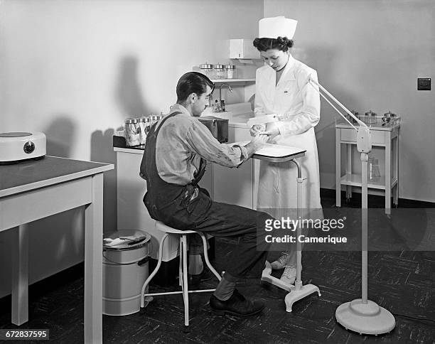 1940s 1950s NURSE EXAMINING INJURY ON HAND OF MAN IN FACTORY INFIRMARY