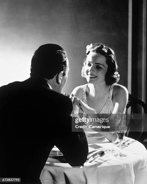 1930s ROMANTIC COUPLE MAN AND WOMAN HOLDING HANDS SEATED AT AN ELEGANT DINNER TABLE