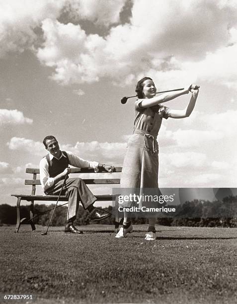 1930s COUPLE PLAYING GOLF MAN ON BENCH COACHING WOMAN SWINGING CLUB