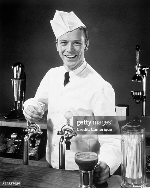 1940s 1950s SMILING YOUNG MAN SODA JERK AT COUNTER SERVING AN ICE CREAM SODA LOOKING AT CAMERA